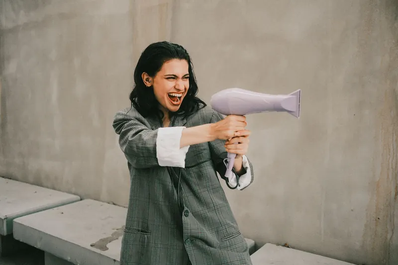 Joyful woman in oversized coat holding a hair dryer, mimicking a playful pose