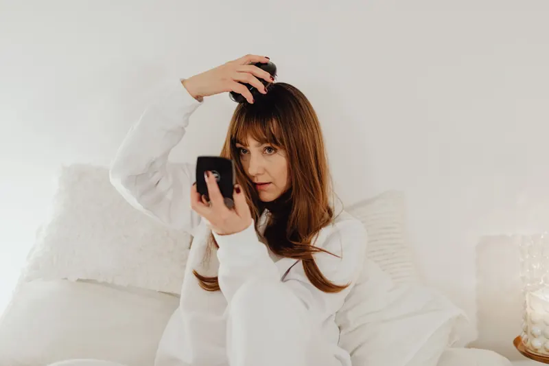 Woman in white attire combs her hair while seated on a bed with soft lighting