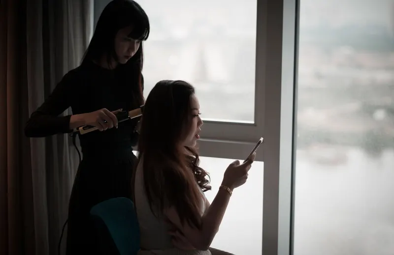 Asian woman curls hair while using a smartphone by a window in dim light